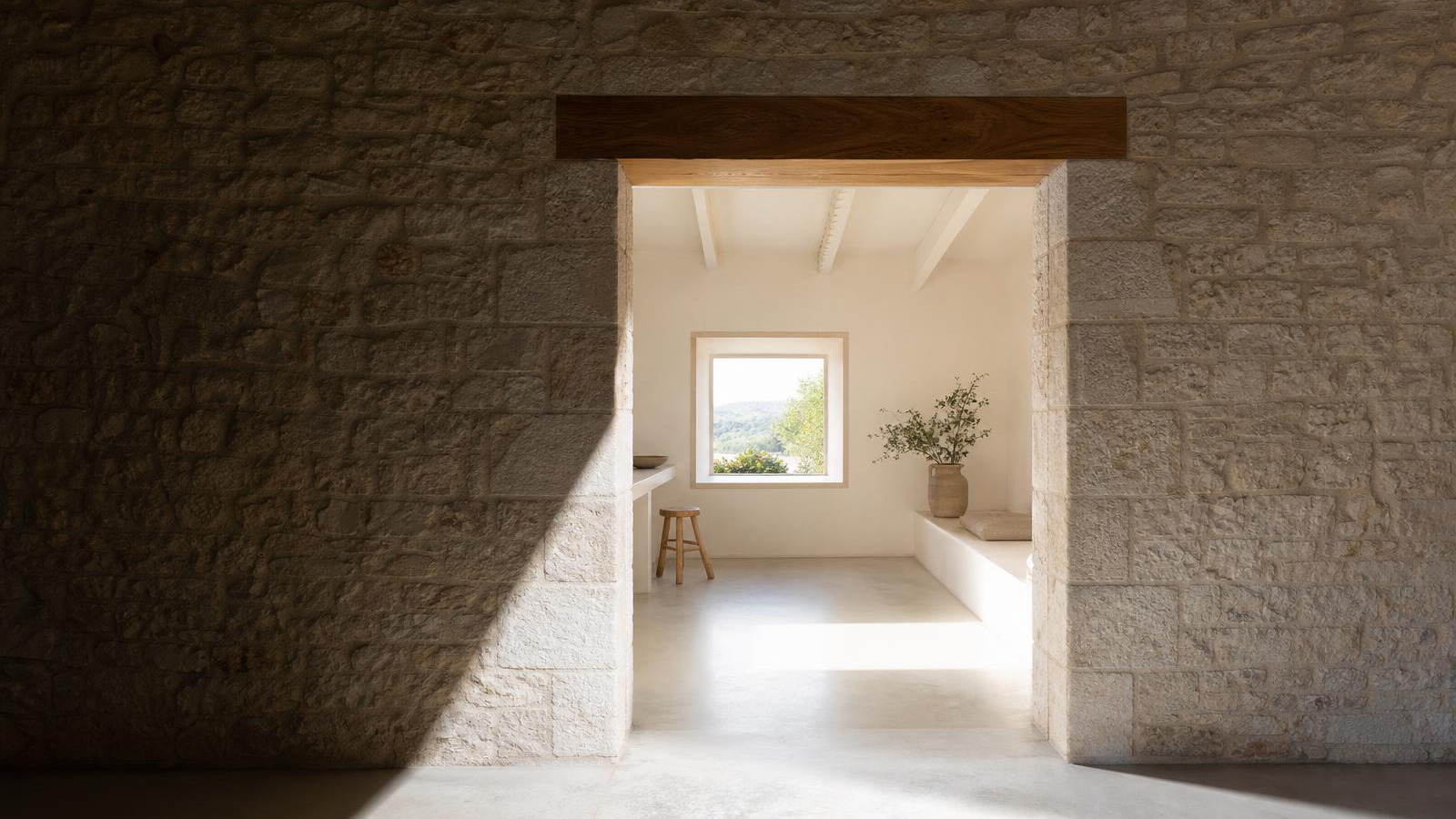 Stone farmhouse renovation in the south of France: a deep shadowed limestone wall beside a newly cut square opening with an oak lintel, bright lime-washed interior beyond.