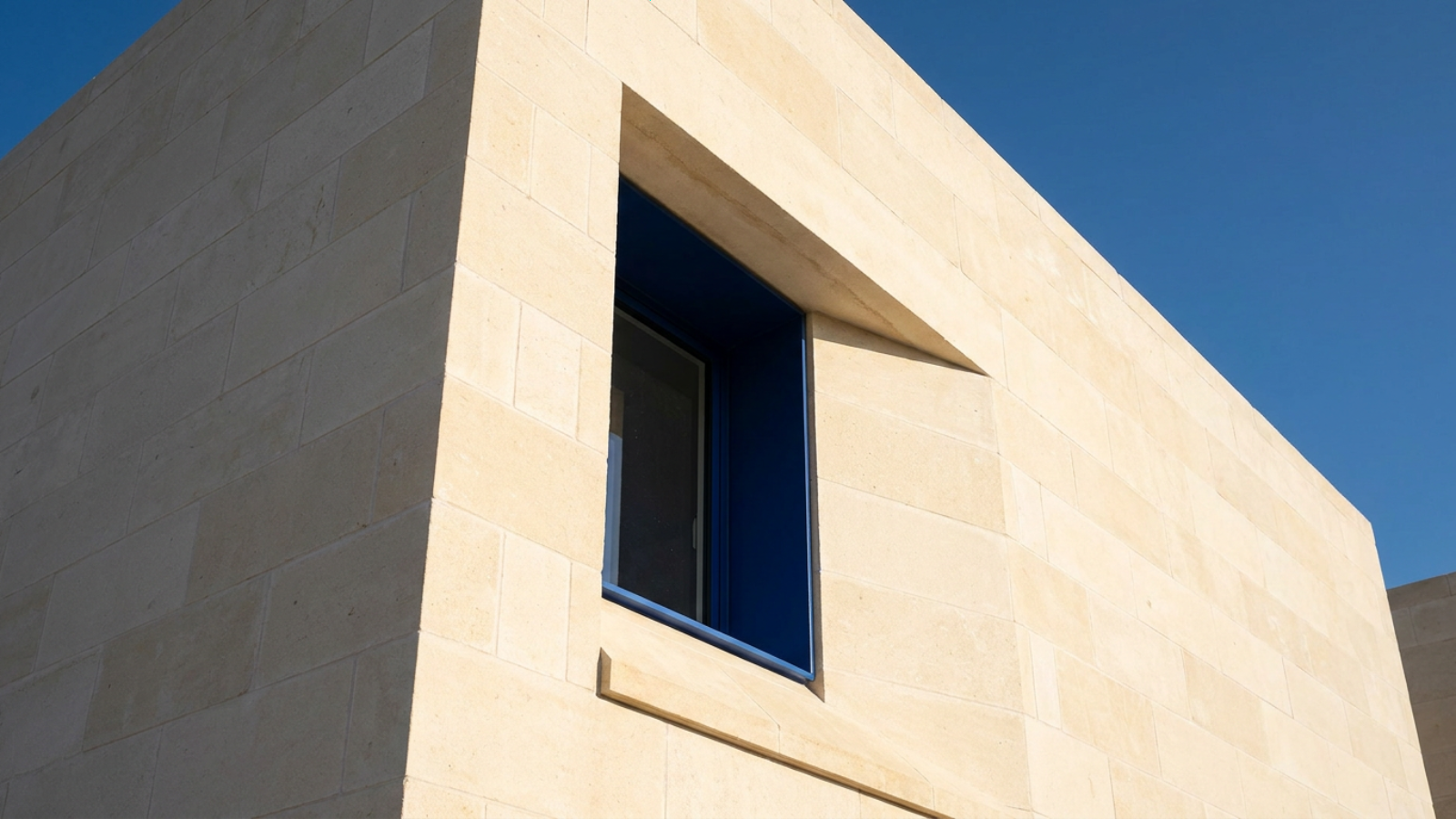 A close-up photograph of a heavy mineral facade showing deep-set window reveals and thick limestone blocks with visible horizontal mortar joints under sharp sunlight.